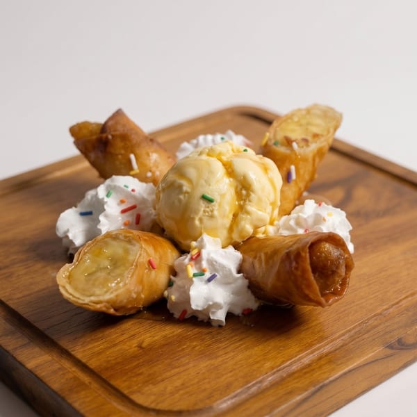 Banana spring rolls with vanilla ice cream, whipped cream, and sprinkles on a wooden board.