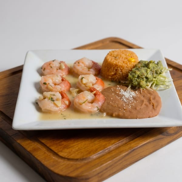 Shrimp scampi with rice, guacamole, and refried beans on a white plate and wooden board.